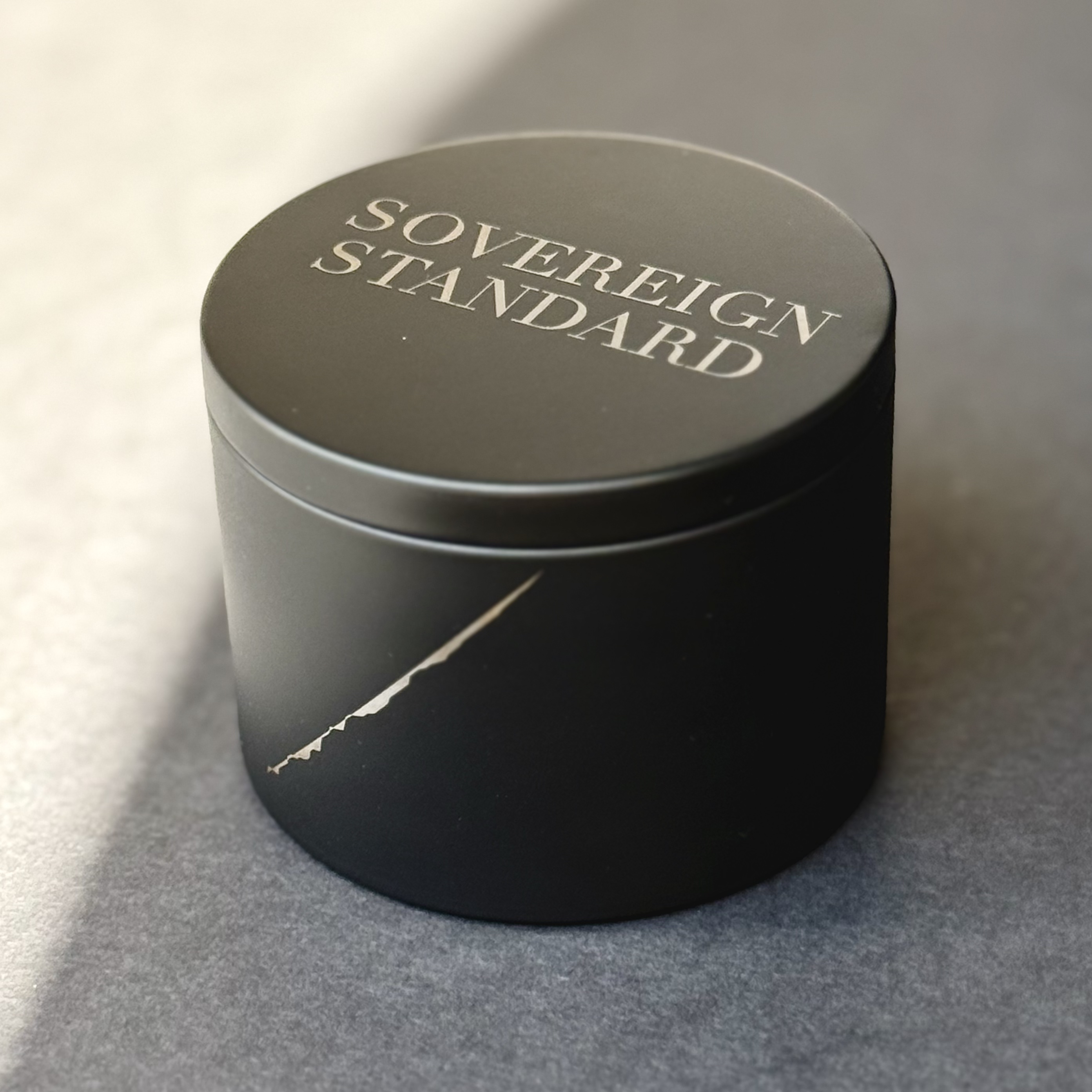 Black Sovereign Standard tea tin vessel resting in warm side light, its engraved wordmark and silver material line emerging softly from shadow